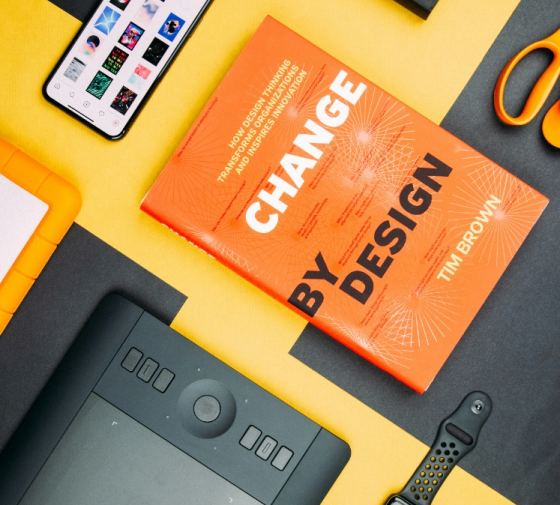 Overhead view of a workspace featuring a book titled "Change by Design" by Tim Brown, a graphic tablet, smartphone, and various office accessories on a yellow and black background.