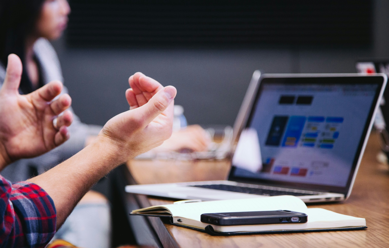 A close-up of a person explaining UX design concepts to colleagues during a collaborative meeting, with a laptop displaying user interface designs on the screen. The scene captures the interaction and brainstorming essential in UX research and design processes.