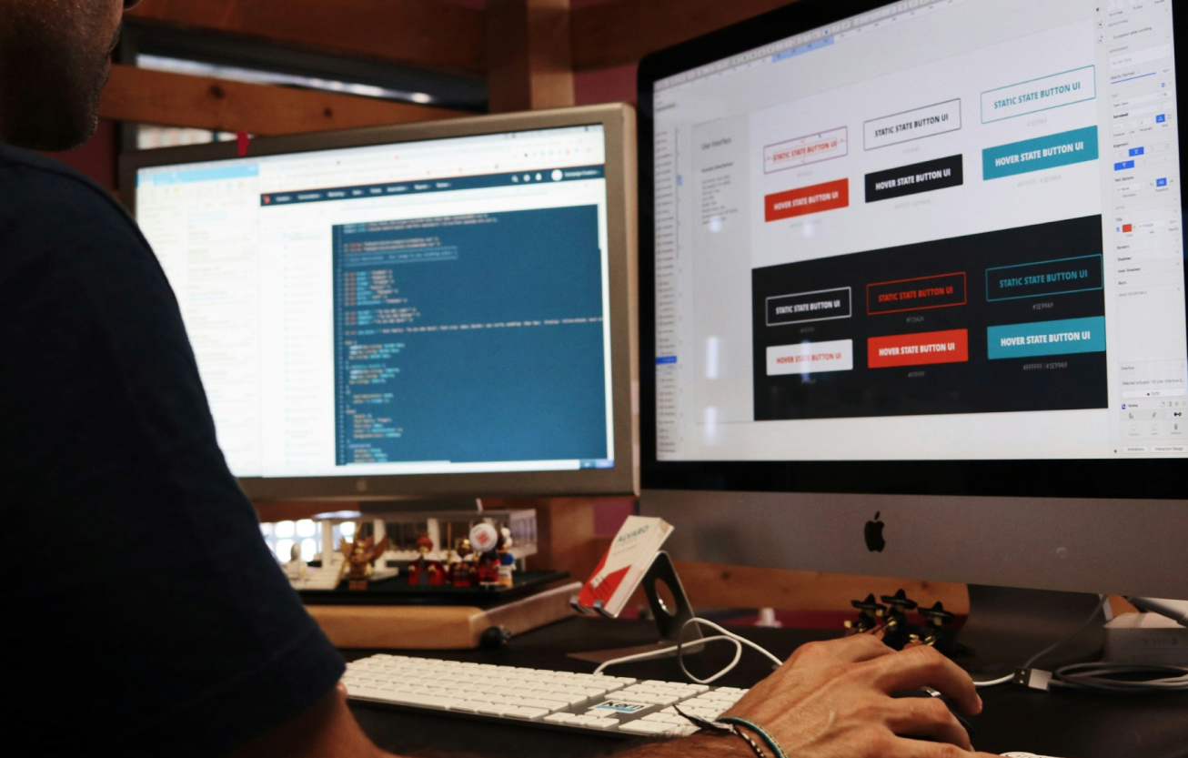 A designer working on interactive prototyping and product design on dual monitors, one displaying UI button variations and the other showing code. The workspace highlights the integration of design and development in creating interactive prototypes for user testing.